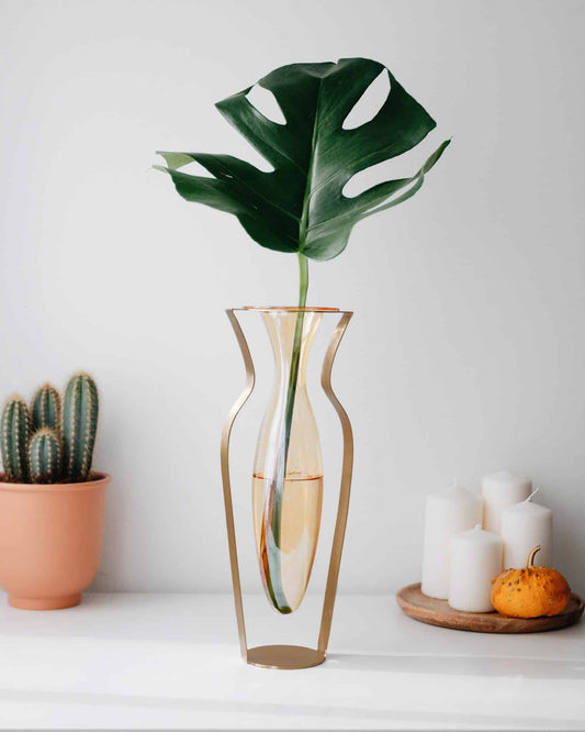 Elegant glasvase med gyldne messingdetaljer og grøn Monstera-blad i moderne boligindretning.